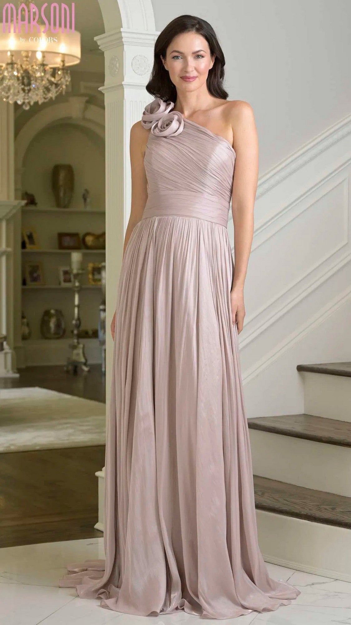 A woman stands indoors near a staircase, gracefully wearing the Marsoni M332 Long Formal Dress in light mauve—a floor-length, one-shoulder chiffon evening gown with a 3D floral embellishment on the strap.
